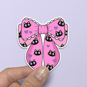 May include: A pink bow-shaped sticker with a repeating pattern of black cat faces and purple hearts. The bow has a scalloped edge and is held by a hand against a light purple background. The sticker is a cute, whimsical design.