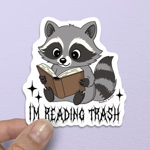 Puede incluir: Una pegatina con un mapache de dibujos animados leyendo un libro. El mapache es gris y blanco con una m&aacute;scara negra y una cola a rayas. El texto "I'M READING TRASH" est&aacute; debajo en una fuente g&oacute;tica.
