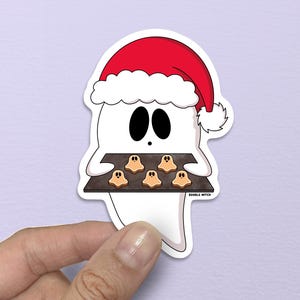 May include: A white ghost sticker wearing a red Santa hat, holding a tray of ghost-shaped cookies. The ghost has black oval eyes and a smiling mouth. The cookies are light brown. The sticker is held against a light purple background.