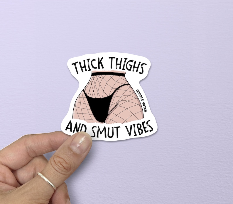 Op de afbeelding: Een witte sticker met de tekst "THICK THIGHS AND SMUT VIBES" en een illustratie van het onderlichaam van een persoon in zwart ondergoed en visnetkousen. De sticker wordt door een hand vastgehouden.