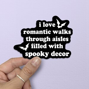 Puede incluir: Una pegatina negra troquelada con texto blanco que dice: "i love romantic walks through aisles filled with spooky decor". El dise&ntilde;o presenta dos siluetas de murci&eacute;lagos blancos, una situada en la parte superior derecha y otra en la inferior izquierda.