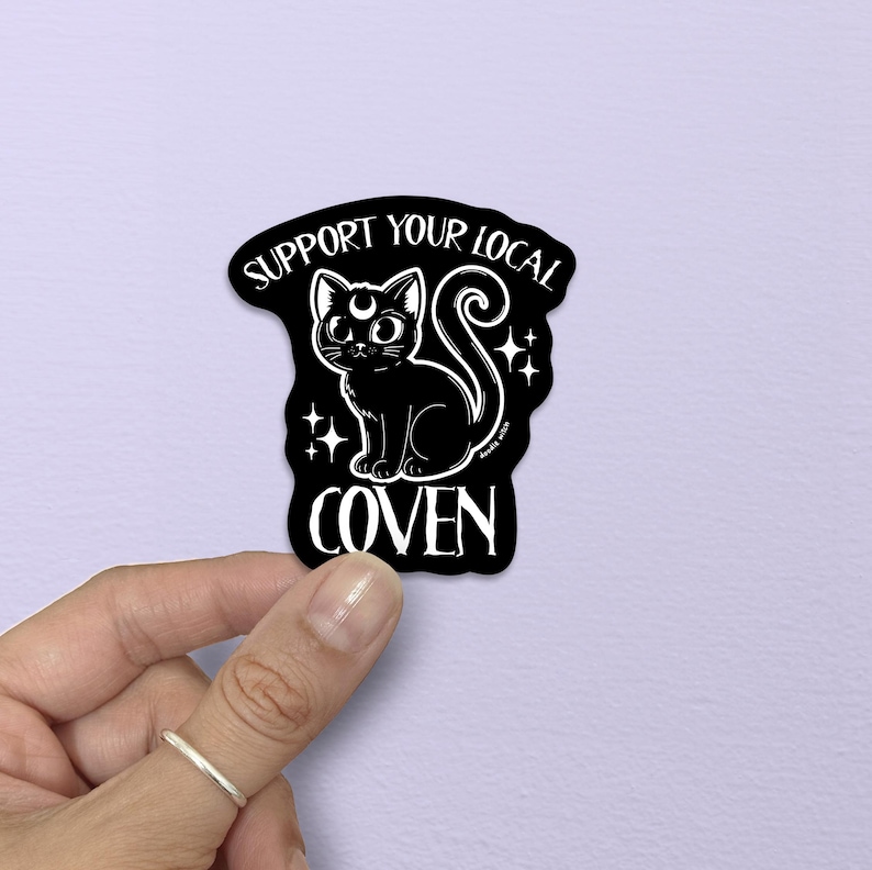 May include: Black sticker with a white illustration of a cat with a crescent moon on its head, surrounded by stars. The text "SUPPORT YOUR LOCAL COVEN" is written in a stylized font.