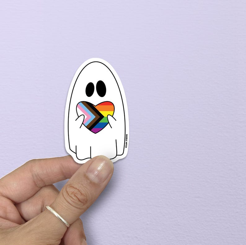 K&ouml;nnte beinhalten: Ein wei&szlig;er Geisteraufkleber mit schwarzen Augen und einem Regenbogenherz. Das Herz zeigt die Farben der Pride-Flagge und der Progress-Pride-Flagge. Der Aufkleber wird von einer Hand vor einem hellvioletten Hintergrund gehalten.