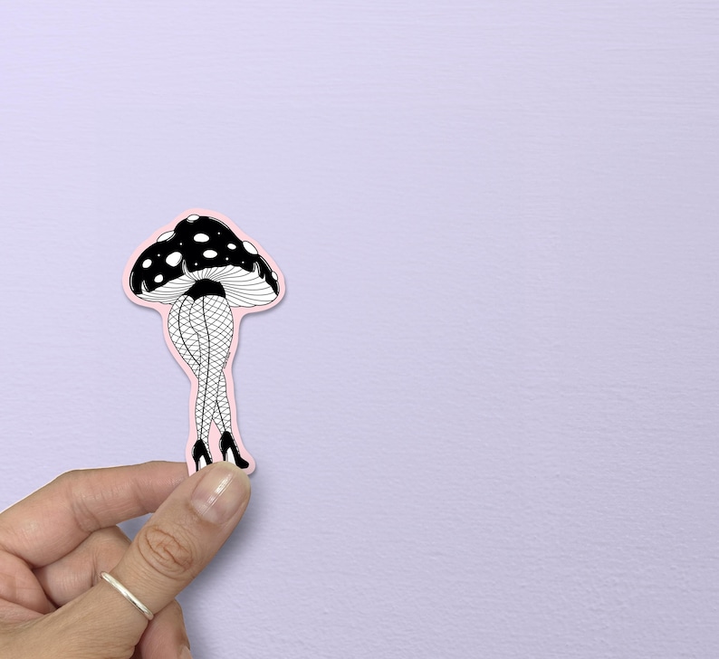May include: A whimsical sticker featuring a black and white mushroom cap with white polka dots. The mushroom has legs clad in fishnet stockings and black high heels, set against a pink outline. The sticker is held by a hand.