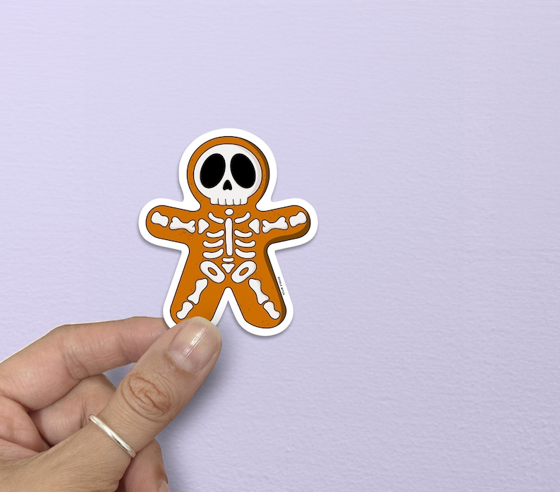 May include: A Halloween-themed sticker in the shape of a gingerbread man, featuring a skeleton design. The sticker is orange with white and black details, held against a light purple background.