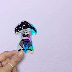 May include: Holographic sticker featuring a mushroom design with a black and white cap and a lingerie-inspired body. The sticker has a rainbow iridescent effect, with a black bra, garter belt, and thigh highs. The sticker is held by a hand.