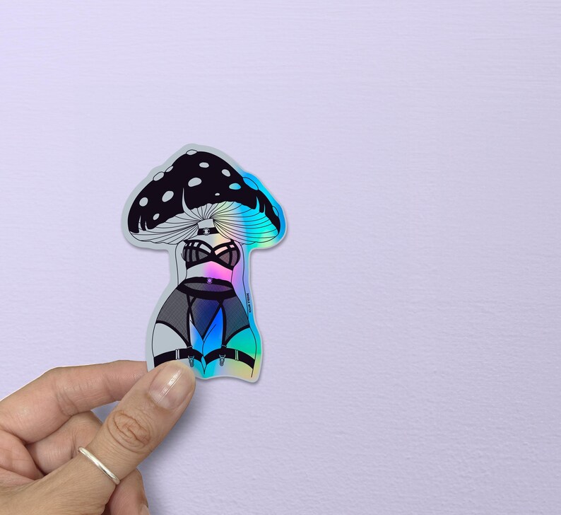 May include: Holographic sticker featuring a mushroom design with a black and white cap and a lingerie-inspired body. The sticker has a rainbow iridescent effect, with a black bra, garter belt, and thigh highs. The sticker is held by a hand.