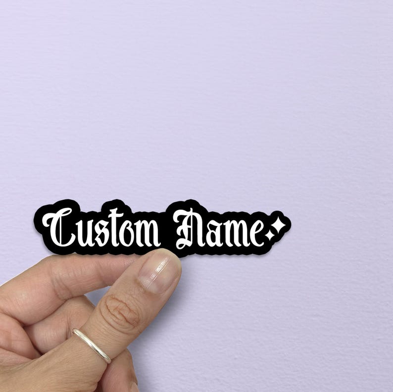 May include: A black sticker with the words "Custom Name" in a white gothic font. The sticker has a black border and is held by a hand with a silver ring. The background is a light purple color.
