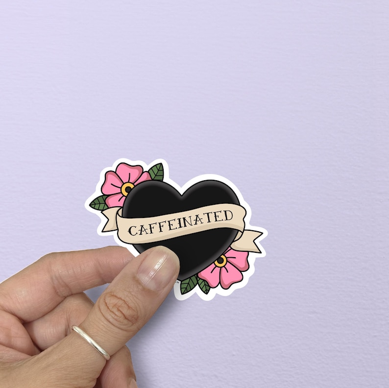 May include: A black heart-shaped sticker with the word "CAFFEINATED" on a banner. The sticker is decorated with pink flowers and green leaves. The sticker is held by a hand.