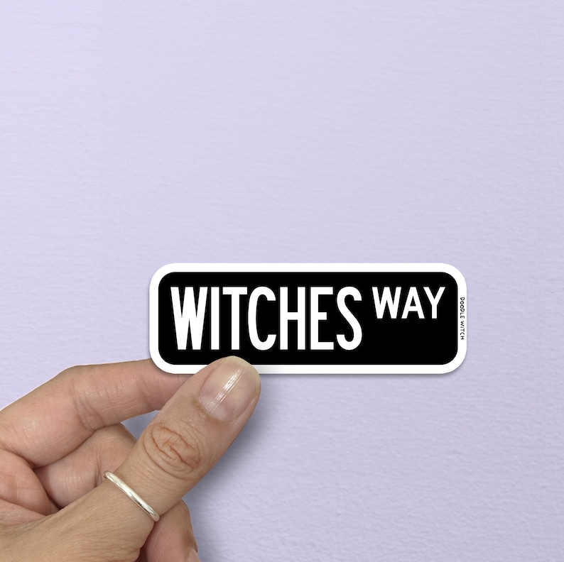 May include: A black and white sticker with the words "WITCHES WAY" in a bold, sans-serif font. The sticker has a white border and is held by a hand against a light purple background. The sticker is rectangular with rounded corners.