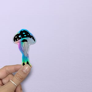May include: A holographic sticker featuring a mushroom design. The mushroom cap is black with white spots, and the stem depicts legs in fishnet stockings and high heels. The sticker has a rainbow iridescent effect.