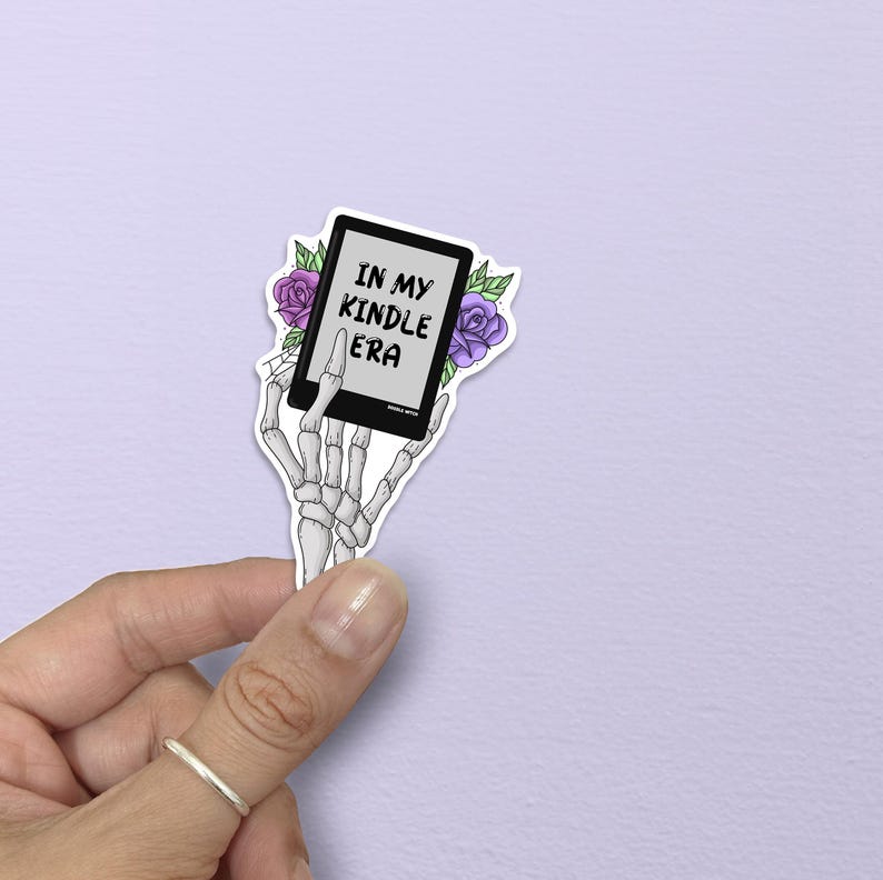 May include: A sticker showing a skeleton hand holding a Kindle. The Kindle screen reads "IN MY KINDLE ERA". The design includes purple roses and green leaves. The sticker has a white border.