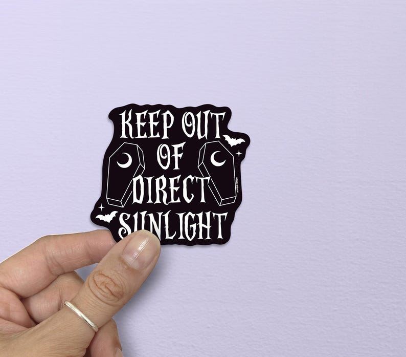 以下が含まれることがあります： 「KEEP OUT OF DIRECT SUNLIGHT」の白い文字が入った黒いステッカー。棺とコウモリのイラストが特徴です。淡い紫色の背景に、手でステッカーを持っています。