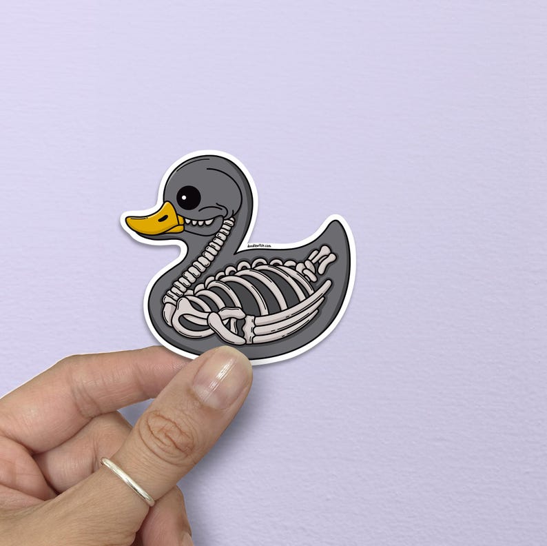 May include: A gray sticker of a rubber duck with a skeleton design. The duck has a yellow beak and a black eye. The skeleton is white and detailed, showing the rib cage and other bones. The sticker is held by a hand.