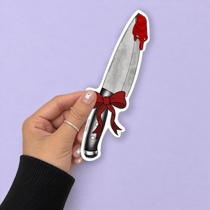 May include: A die-cut sticker featuring a silver kitchen knife with a black handle, tied with a dark red ribbon. The blade has a drip of bright red liquid at the tip. The text "DOODLE WITCH" is printed in small black letters on the knife handle.
