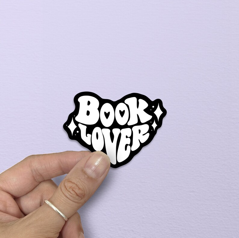 May include: A black and white sticker with the words "BOOK LOVER" in a retro font. The sticker is heart-shaped with small hearts and diamond shapes. The sticker is held by a hand against a light purple background.