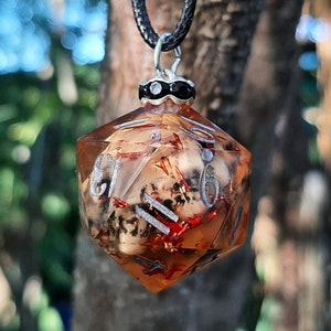 Peut inclure: Un pendentif en résine transparente en forme de dé avec un crâne et des paillettes rouges et blanches à l'intérieur. Le dé est un dé à 20 faces avec les chiffres de 1 à 20 visibles. Le pendentif est suspendu à un cordon noir.