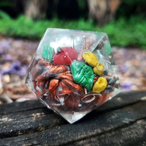 May include: A clear resin dice with a six-sided shape. The dice is filled with colorful, small, resin pieces, including red, orange, yellow, and green. The dice is sitting on a dark brown wooden surface.
