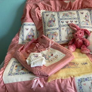May include: A pink baby bedding set with embroidered animal and heart designs. The set includes a quilt, pillow, blanket, tote bag, and a small stuffed bunny. The quilt and pillow feature squares with various animals. A small white shirt is also included.