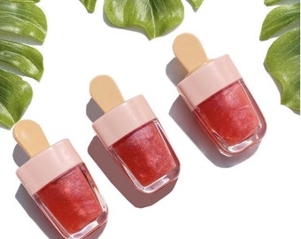 May include: Three popsicle-shaped lip glosses with a pink and clear plastic container and a wooden stick. The lip gloss is a shimmery red color.