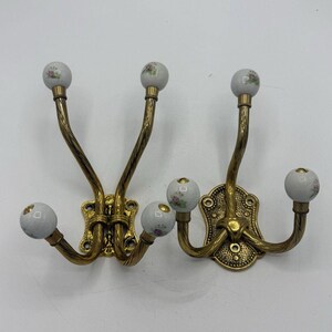 May include: Two wall-mounted coat hooks made of antique brass-toned metal with twisted arms. Each hook features white ceramic ball finials decorated with small pink and green floral patterns. The mounting plates have a textured, ornate design.