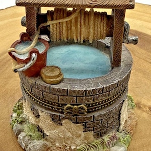 Pode incluir: Estatueta em miniatura de um poço dos desejos com um pássaro azul empoleirado no topo. O poço é feito de tijolo e madeira, com um balde vermelho e uma pequena moeda. O poço mede aproximadamente 10 cm de altura.