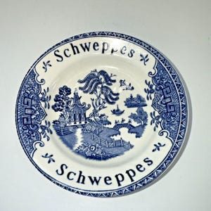 May include: A white and blue ceramic plate with a landscape design. The plate features a blue and white Asian-inspired scene with a pagoda, trees, and a bridge. The word "Schweppes" is printed in blue.