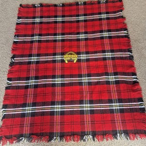 May include: A red, black, and white tartan blanket with a fringed edge. The blanket features a repeating pattern of intersecting lines. A gold emblem with the word "Horseshoe" is in the centre.