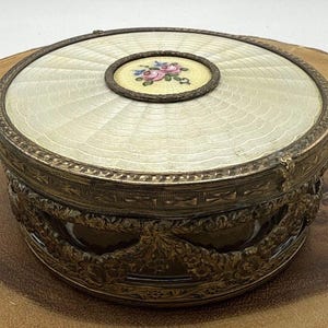 Pode incluir: Uma caixa de joias redonda e antiga com uma tampa creme com um padrão radial. A tampa tem um medalhão central com um desenho floral em rosa, azul e verde. A caixa tem uma moldura e base de metal decorativas e ornamentadas.