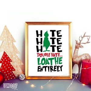 May include: A framed print featuring a green Grinch silhouette and the text "Hate Hate Hate Double Hate... Loathe Entirely". The print is on a white background and has a gold frame. The print is displayed on a table with a Christmas tree and a gold reindeer figurine.