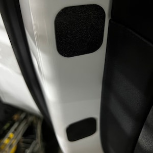 May include: Close-up of a white car door interior with two black sound dampening pads. The pads are rectangular and have a textured surface.
