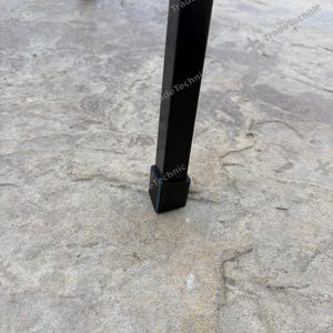 May include: A black metal furniture leg with a square cross-section and a black plastic protective cap at the base. The leg is positioned vertically, and the text TradeTechnic is visible as a watermark across the image.