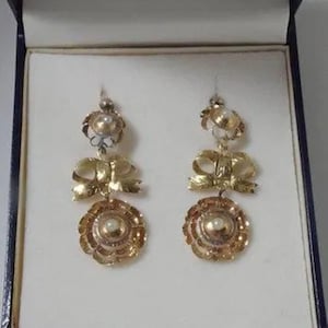 Könnte beinhalten: Ein Paar goldene Ohrringe mit Perlenakzenten. Jeder Ohrring hat einen runden Anhänger mit einer Perle in der Mitte, umgeben von einem zarten Blumenmuster. Oberhalb des Anhängers befinden sich zwei Schleifen, ebenfalls aus Gold. Die Ohrringe werden in einer blauen Samtbox präsentiert.