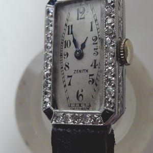 May include: A vintage Zenith wristwatch with a rectangular face, surrounded by a diamond-studded bezel. The watch has a silver dial with black numerals and hands, and a black leather strap. The watch face reads "Zenith" and "Swiss Made".