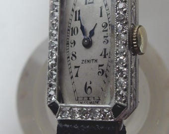 May include: A vintage Zenith wristwatch with a rectangular face, surrounded by a diamond-studded bezel. The watch has a silver dial with black numerals and hands, and a black leather strap. The watch face reads "Zenith" and "Swiss Made".