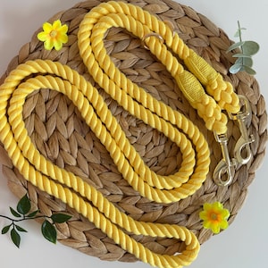 Könnte beinhalten: Eine gelbe Hundeleine aus Baumwollseil mit zwei goldenen Metallclips. Die Leine ist auf einer geflochtenen Strohoberfläche aufgerollt.