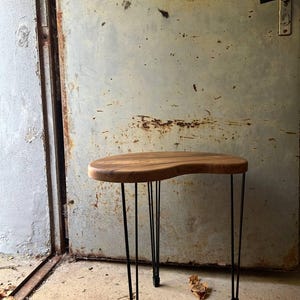 Könnte beinhalten: Ein nierenförmiger Beistelltisch mit einer Tischplatte aus Naturholz mit sichtbarer Maserung und abgerundeten Kanten. Der Tisch steht auf vier dünnen, schwarzen Haarnadelbeinen aus Metall, die senkrecht zum Boden verlaufen.