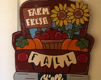 May include: A wooden cutout of a red pickup truck with a banner that says "Fall Y'all". The truck bed is filled with pumpkins, apples, and sunflowers. The truck has a sign that says "Farm Fresh".
