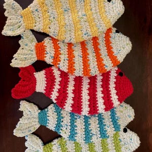Puede incluir: Cuatro peces de ganchillo coloridos en amarillo, naranja, rojo y azul con rayas blancas y aletas caudales. Cada pez tiene un ojo negro. Los peces están apilados uno encima del otro.