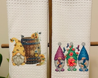 Peut inclure: Deux torchons de cuisine blancs avec une texture en nid d'abeille. Le torchon de gauche représente un gnome avec un chapeau jaune et un tonneau portant l'inscription "Honey". Le torchon de droite représente trois gnomes avec des chapeaux violets, verts et bleus, chacun tenant un colibri.