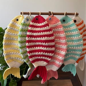 May include: A collection of six colorful, crocheted fish hanging from a wooden rod. The fish feature various colors, including yellow, green, red, pink, teal, and orange, with white accents. Each fish has a unique pattern and a tail.