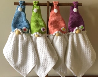 Peut inclure: Quatre torchons de cuisine en forme de gnome crochetés suspendus à un support en bois. Les gnomes portent des chapeaux bleus, verts, roses et violets avec des fleurs blanches sur le bord. Les torchons sont blancs avec une texture gaufré.