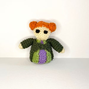 Puede incluir: Muñeca de crochet con pelo naranja, ropa verde y morada y ojos negros.