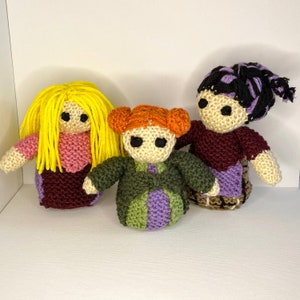May include: Three crocheted dolls with different coloured hair and clothing. The doll on the left has yellow hair and a pink and burgundy dress. The doll in the middle has orange hair and a green and purple dress. The doll on the right has black hair and a burgundy and purple dress.