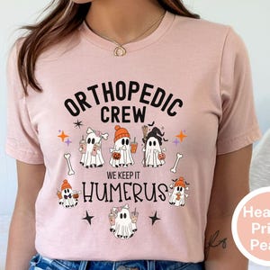 May include: A pink t-shirt with the text "Orthopedic Crew We Keep It Humerus" and a graphic of five ghosts holding drinks and sweets. The ghosts are wearing hats and have different facial expressions.