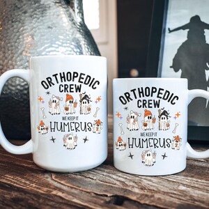 May include: Two white ceramic coffee mugs with a Halloween-themed design. The mugs feature a group of ghosts dressed as orthopedic surgeons with the text "Orthopedic Crew We Keep It Humerus".