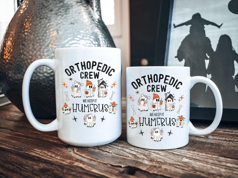 May include: Two white ceramic coffee mugs with a Halloween-themed design. The mugs feature a group of ghosts dressed as orthopedic surgeons with the text "Orthopedic Crew We Keep It Humerus".
