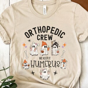 May include: A tan t-shirt with the text "Orthopedic Crew" and "We Keep It Humerus" in black. The shirt features a Halloween theme with five cartoon ghosts holding coffee cups and pumpkins.