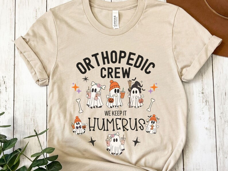 May include: A tan t-shirt with the text "Orthopedic Crew" and "We Keep It Humerus" in black. The shirt features a Halloween theme with five cartoon ghosts holding coffee cups and pumpkins.