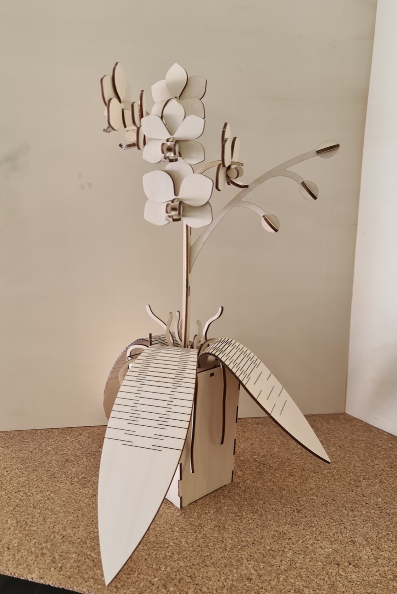 Peut inclure: Une sculpture de fleur d'orchid&eacute;e en bois blanc avec un design d&eacute;taill&eacute;. La fleur est dans un pot en bois avec des feuilles.